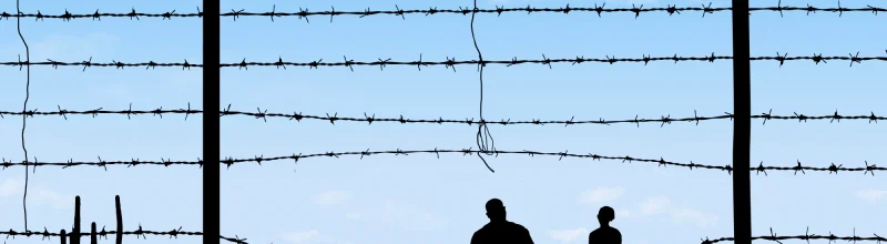 Silhouette of two people crossing through a barbed fence.